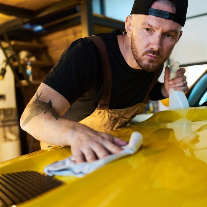 Mann mit Kappe reinigt aufmerksam die Motorhaube eines gelben Autos.